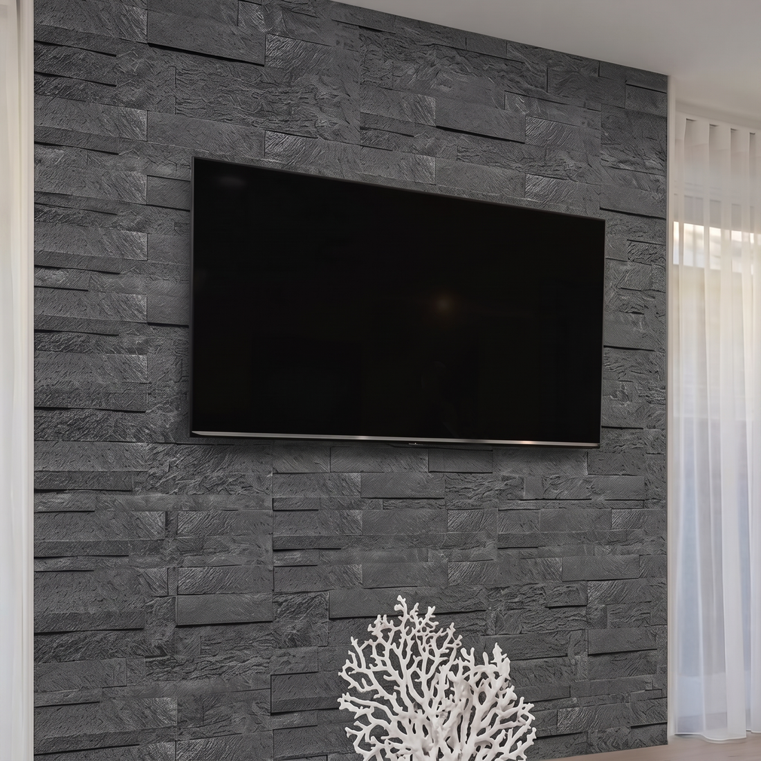La pared de la televisión cambia por completo el salón con los paneles de piedra apilada autoadhesivos Galaxia: piedra 100 % natural, sin pegamento y con una instalación rápida y sencilla, cuyas piezas rugosas en tonos carbón crean un relieve intenso y profundo, y junto al coral blanco y las cortinas claras, el espacio se siente más elegante y con más carácter.